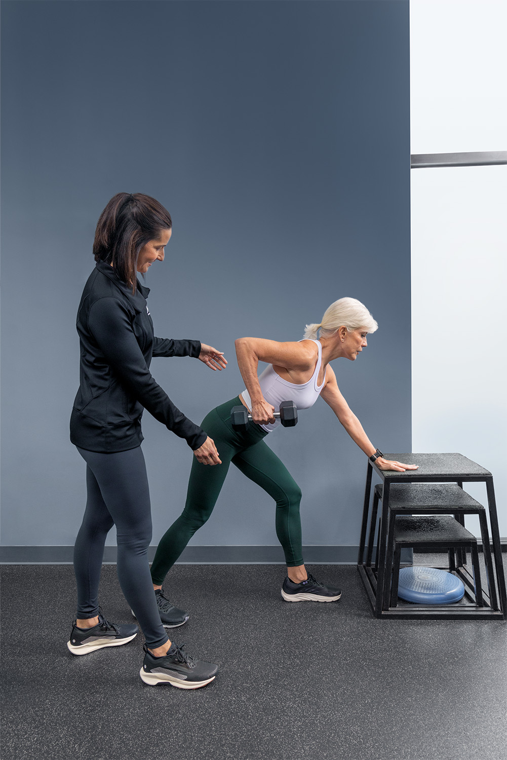 Fitness Together client performing a dumbbell row with her trainer spotting — representing the brand promise 'We're In This Together'